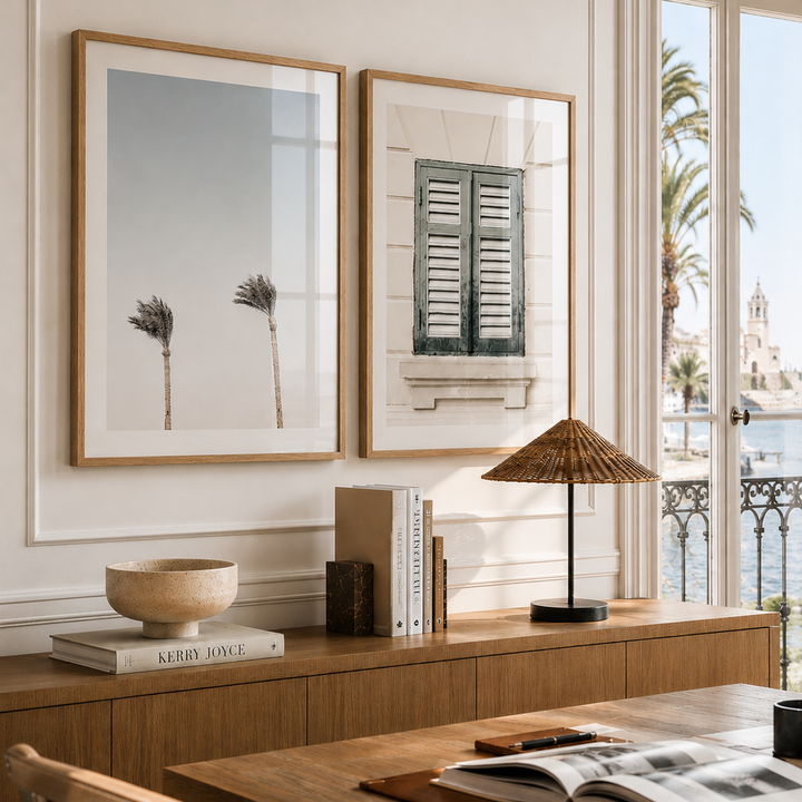 Two framed art prints with light wood frames hang on a cream wall above a wooden console table in a bright, modern room. The left print shows two palm trees, and the right print shows a white building facade with green shutters.