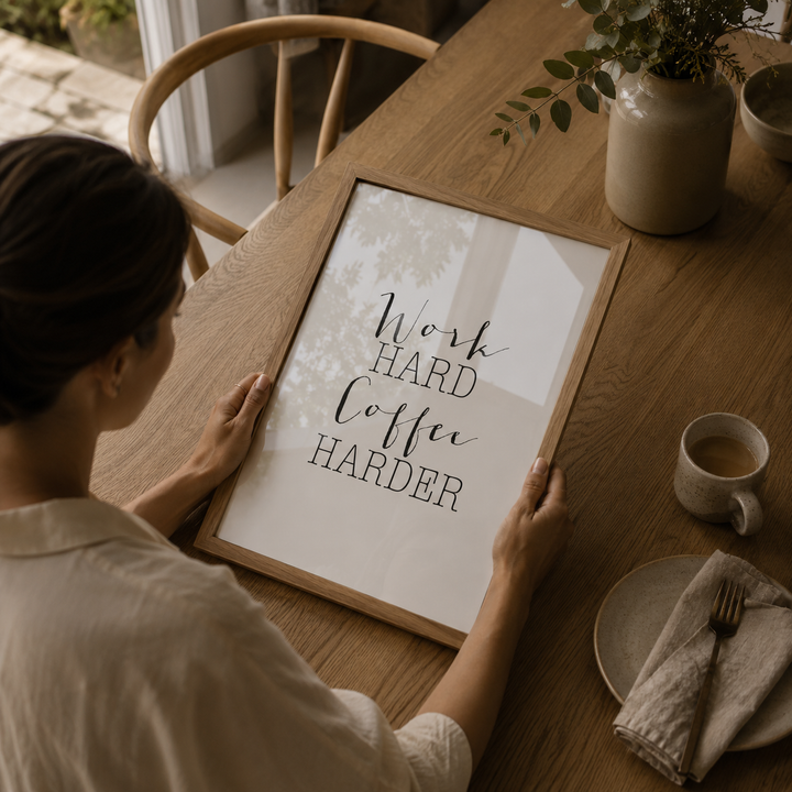 Person holding a framed motivational poster on a wooden table with a cup of coffee and a plant.