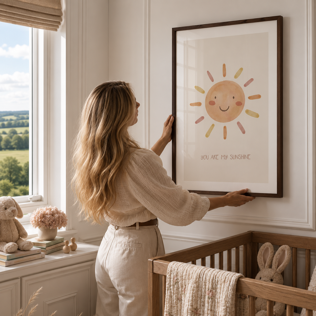 A woman in a cream-colored outfit hangs a framed nursery art print on a white wall in a bright, modern nursery. The print features a cute, smiling sun with pastel-colored rays and the text 'YOU ARE MY SUNSHINE'.