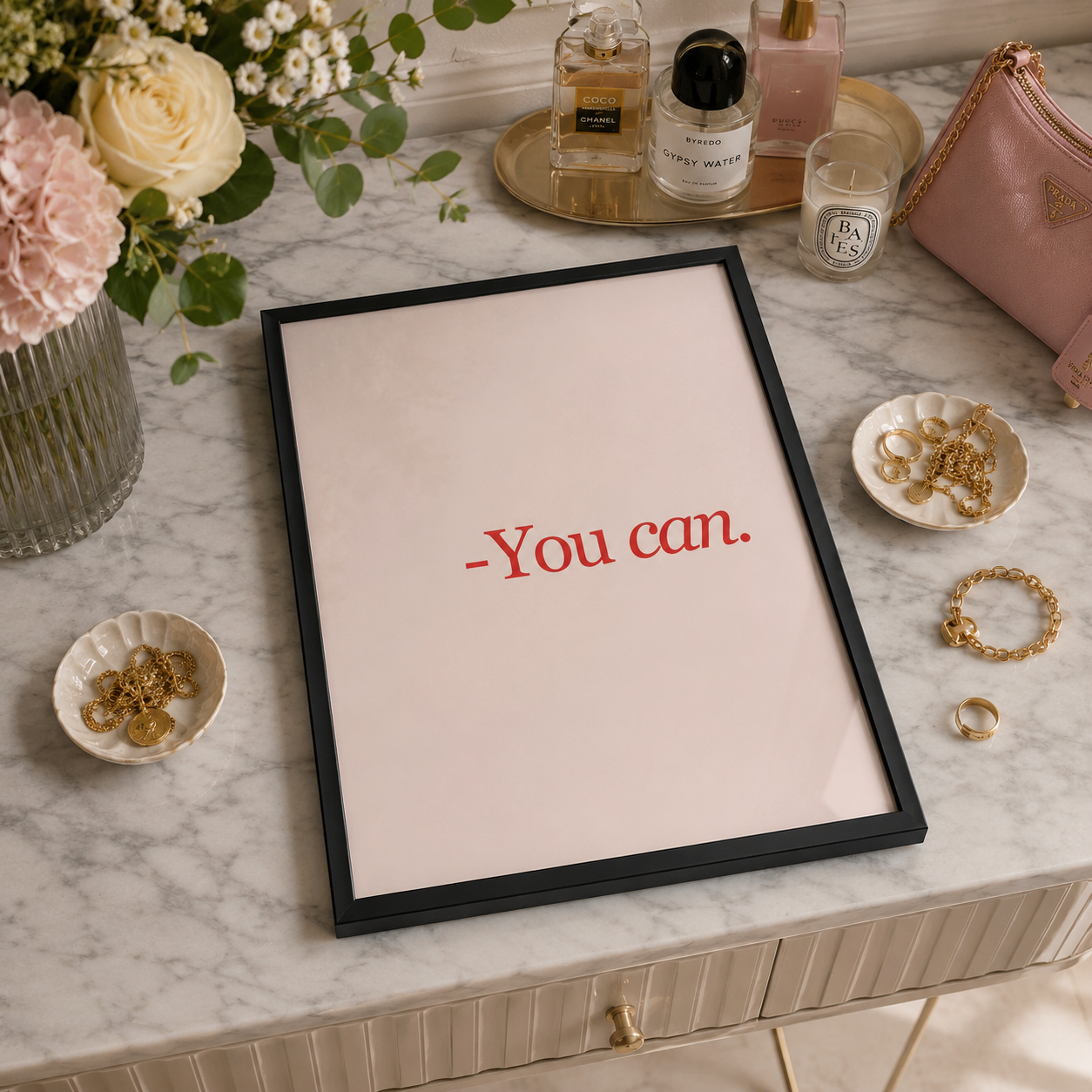 A framed typographic wall art print reading “-You can.” in red serif text on a pale blush background, set in a black frame on a marble surface.