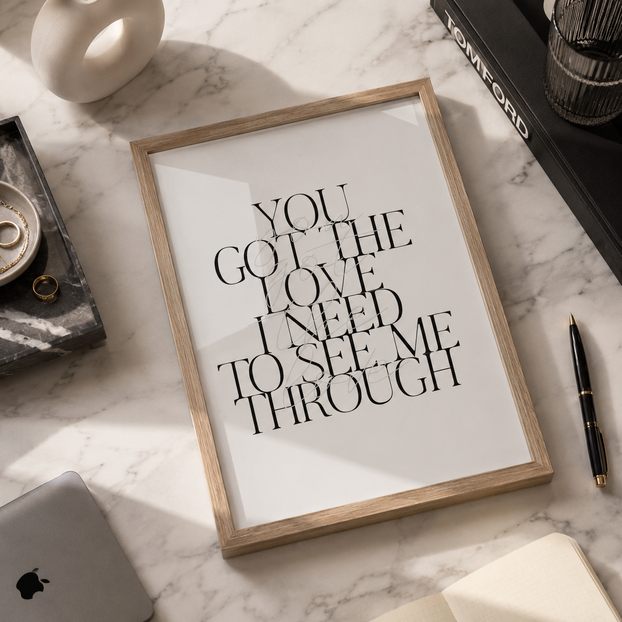 A framed typographic wall art print reading 'YOU GOT THE LOVE I NEED TO SEE ME THROUGH' in black serif text on a white background, set in a light wood frame on a marble surface with styled desk props around it.