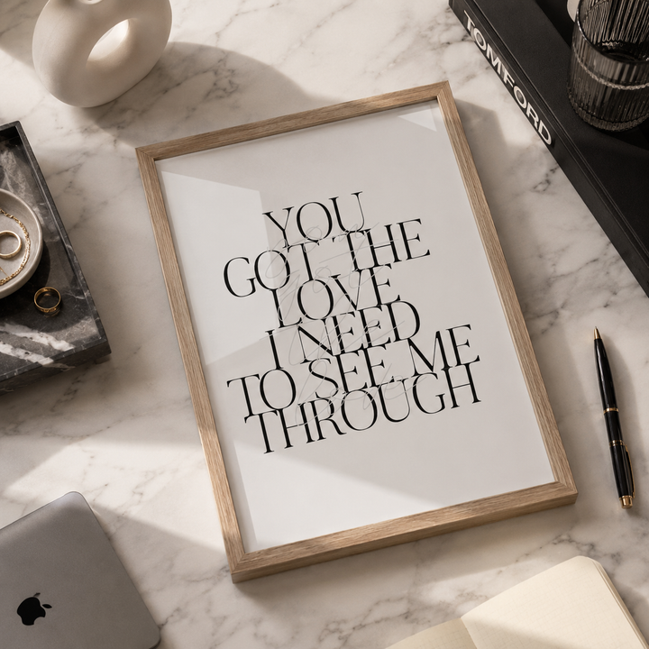 A framed typographic wall art print reading 'YOU GOT THE LOVE I NEED TO SEE ME THROUGH' in black serif text on a white background, set in a light wood frame on a marble surface with styled desk props around it.