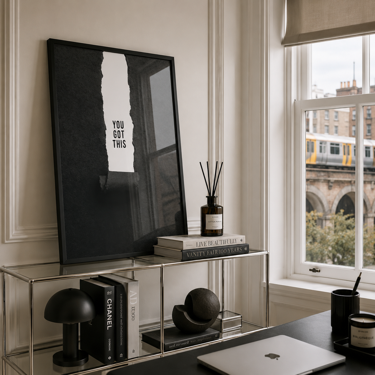 A framed black and white art print with the text 'YOU GOT THIS' leaning on a glass shelving unit in a modern, stylish room with a window overlooking a city scene.