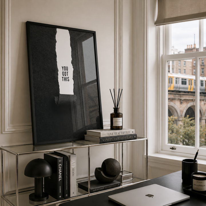 A framed black and white art print with the text 'YOU GOT THIS' leaning on a glass shelving unit in a modern, stylish room with a window overlooking a city scene.