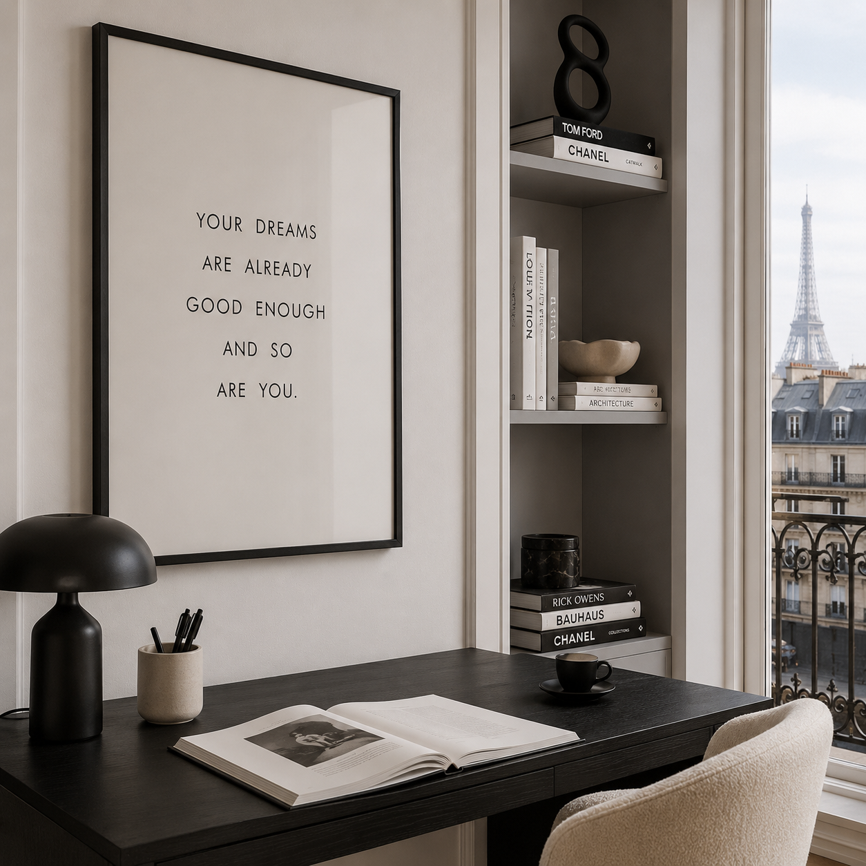 A framed minimalist wall art print with black text on a white background hangs above a dark desk in a styled home office. The room includes a black desk lamp, an open book, a cup, a built-in shelf with books and decor, and a window view of the Eiffel Tower in Paris.