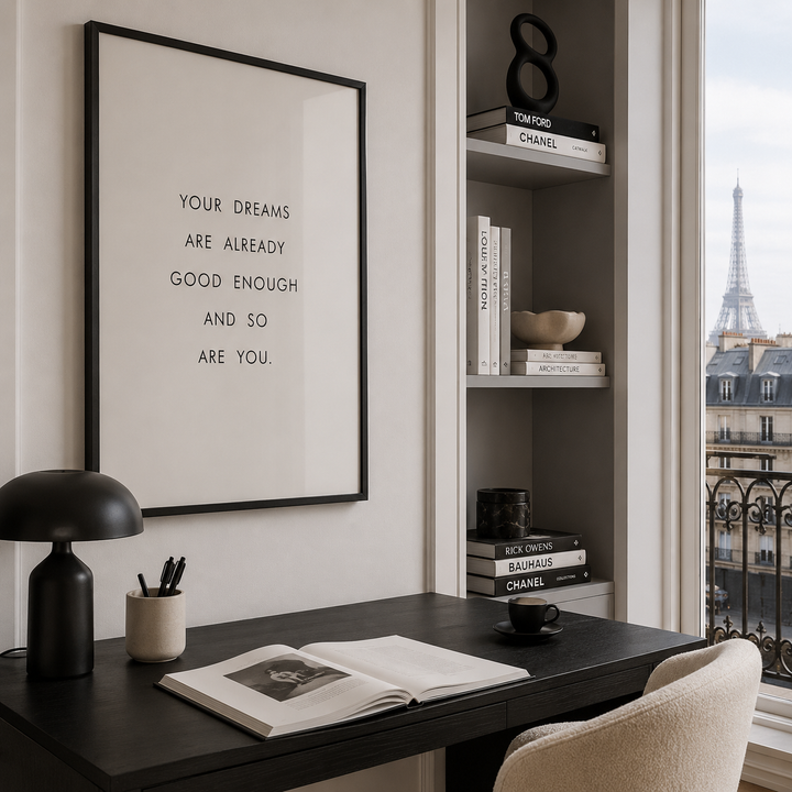A framed minimalist wall art print with black text on a white background hangs above a dark desk in a styled home office. The room includes a black desk lamp, an open book, a cup, a built-in shelf with books and decor, and a window view of the Eiffel Tower in Paris.