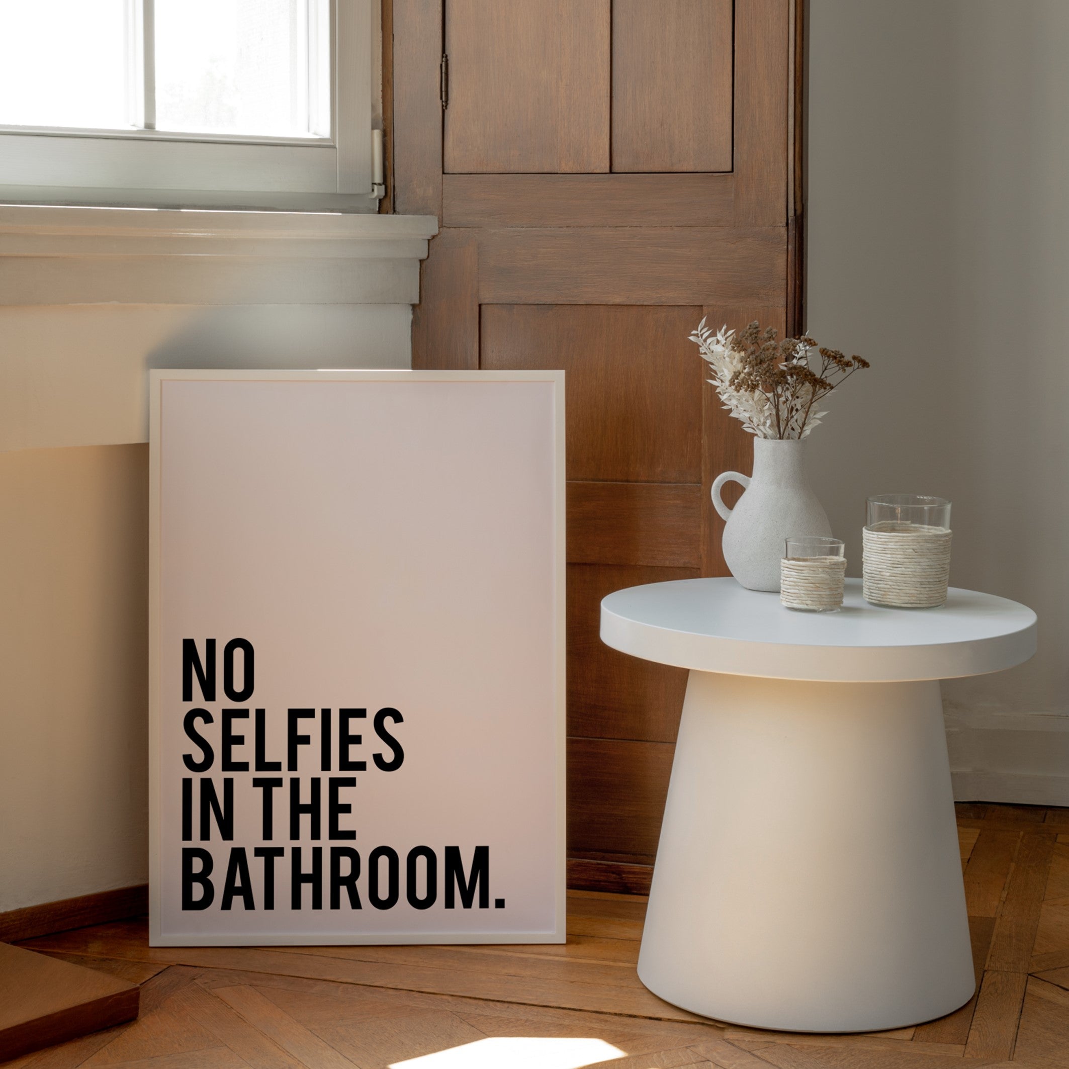Framed poster withe the text "No Selfies In The Bathroom" in black and white resting on the floor of a scandinavian bathroom 